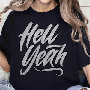 May include: A black short-sleeved t-shirt featuring the text "Hell Yeah" printed in a distressed, light gray, cursive font across the chest. The shirt has a crew neckline and a relaxed fit, with the graphic centered on the front of the garment.