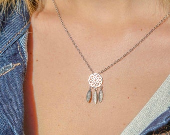 May include: A silver-colored necklace featuring a dreamcatcher pendant with three dangling feather charms. The necklace is worn over a light-colored top and a denim jacket.