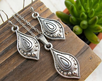 May include: A silver-toned jewelry set featuring a pendant necklace and matching earrings. The pendant and earrings have a teardrop shape with a detailed, textured design. The set is displayed on a wooden surface.