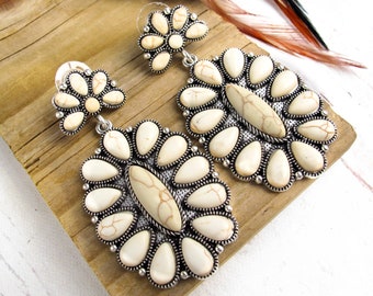 May include: A pair of statement earrings with a floral design. The earrings feature off-white teardrop-shaped stones set in a silver-toned metal. The earrings are displayed on a wooden surface with feathers in the background.