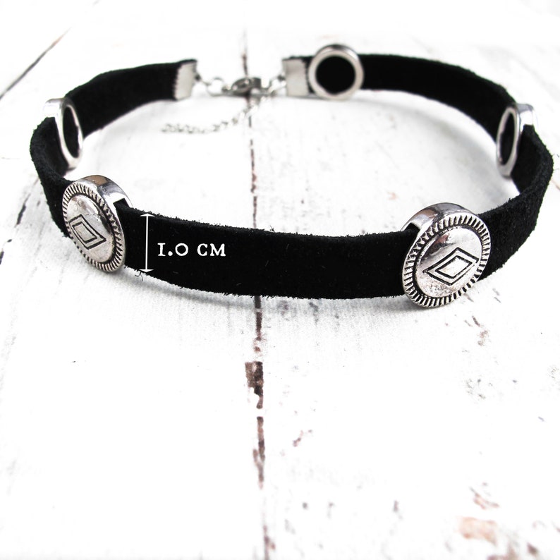 May include: Black suede choker necklace with silver-toned metal accents. The necklace features four round charms with a diamond-shaped design. The choker is 1.0 inches wide.
