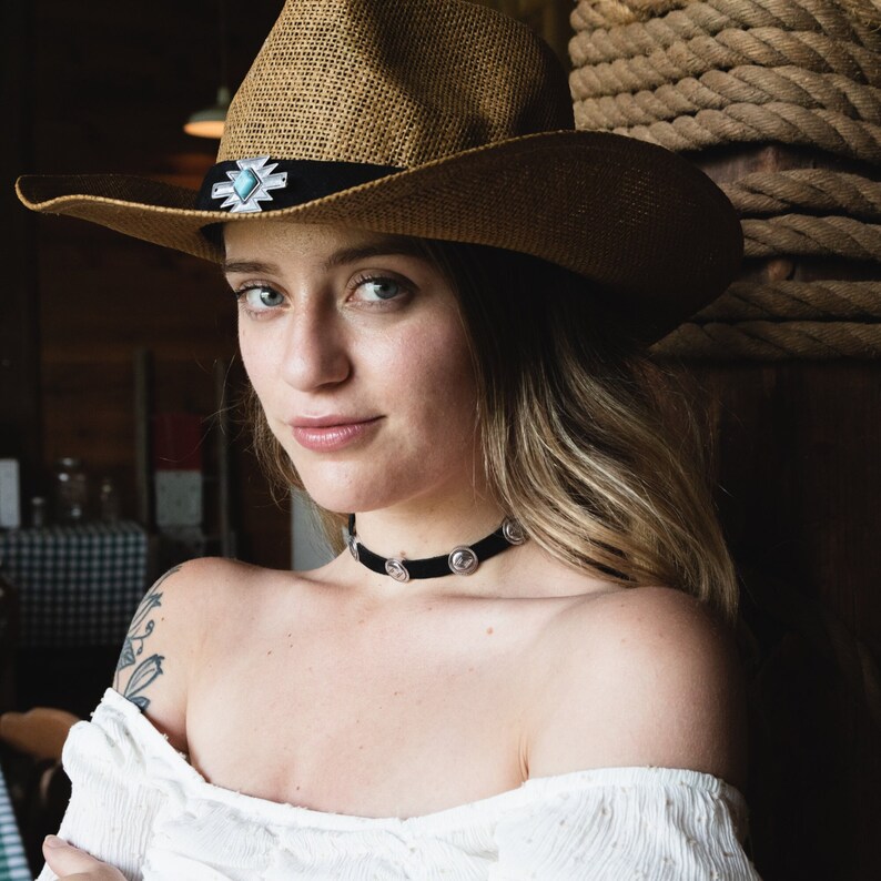 May include: A woman wearing a brown straw cowboy hat with a silver and turquoise concho. She is wearing a black choker necklace with silver accents. The woman has long brown hair and is looking at the camera.