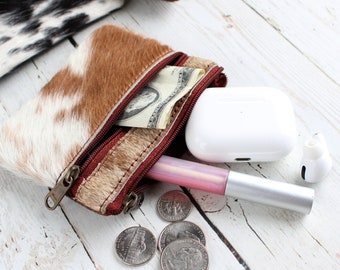 May include: A small brown and white cowhide coin purse with a zipper closure. The purse is open and has a dollar bill inside. There are also a few US quarters, a pink lip gloss, and a white wireless earbud case on a white wooden surface.