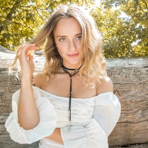 May include: A woman with long blonde hair wearing a white off-the-shoulder top with a black choker necklace and a silver pendant.