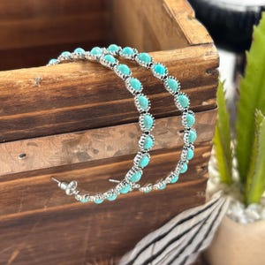 May include: Silver-toned hoop earrings with turquoise-colored stones. The earrings are set in a row of small, round stones. The earrings are displayed on a wooden surface, with a feather and a plant in the background.