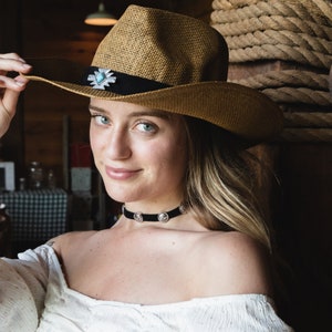 May include: A woman wearing a brown straw cowboy hat with a silver and turquoise concho. She is wearing a black choker necklace with silver accents and a white off-the-shoulder top.
