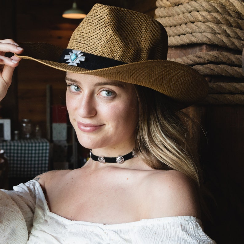 May include: A woman wearing a brown straw cowboy hat with a silver and turquoise concho. She is wearing a black choker necklace with silver accents and a white off-the-shoulder top.