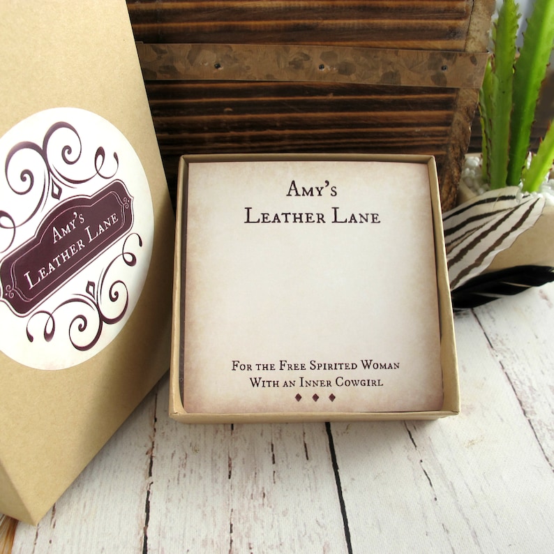 May include: A brown cardboard box with a white circle sticker that says "Amy's Leather Lane". The box is open and inside is a smaller box with a white background and brown text that says "Amy's Leather Lane" and "For the Free Spirited Woman With an Inner Cowgirl".