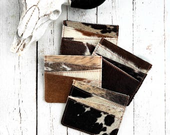 May include: A collection of rectangular card holders crafted from cowhide leather, featuring various natural patterns in shades of brown, black, white, and tan. Each holder displays distinct hair-on-hide textures and stitched edges.