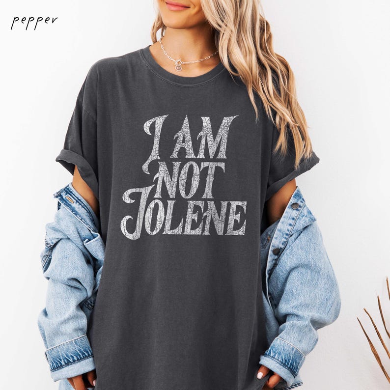 Pu&ograve; includere: T-shirt grigio scuro con la scritta "I AM NOT JOLENE" in un carattere bianco invecchiato. La maglietta ha le maniche arrotolate ed &egrave; indossata con una giacca di jeans azzurra. La maglietta &egrave; uno stile casual e oversize.