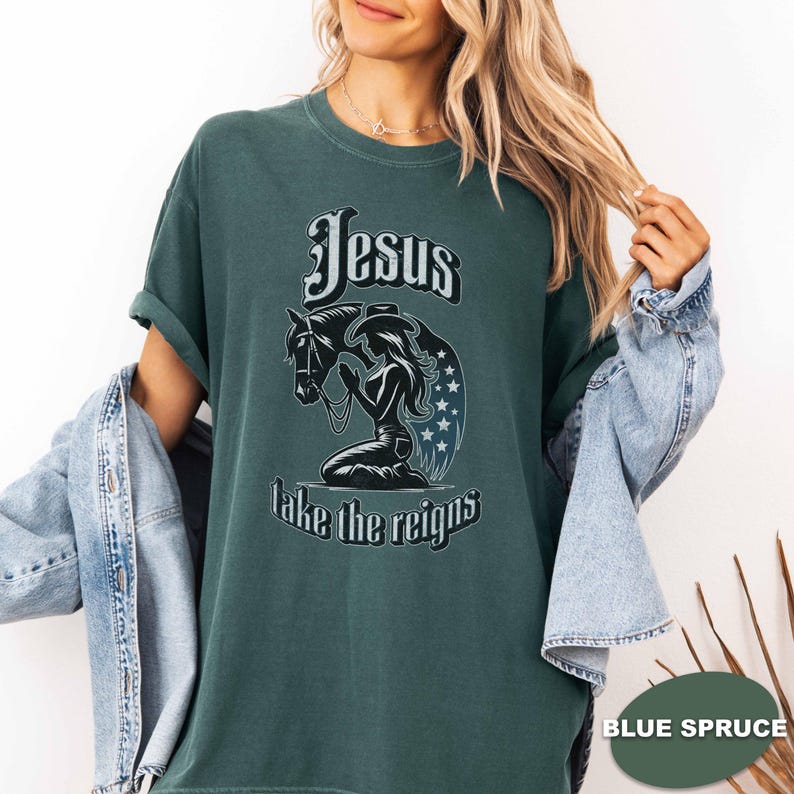 May include: A dark teal t-shirt featuring the text "Jesus take the reigns" above a graphic of a horse and a kneeling woman. The woman is wearing a cowboy hat and praying. The horse has a bridle. The design includes stars.