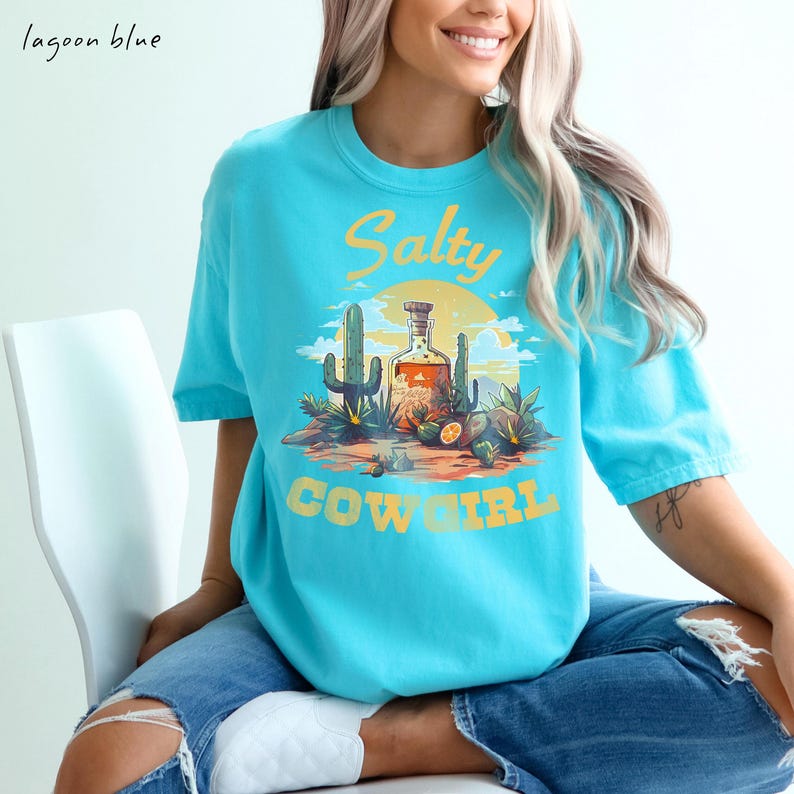 K&ouml;nnte beinhalten: Ein lagunenblaues T-Shirt mit einem W&uuml;sten-Grafikdesign, das Kakteen, eine Flasche und den gelben Schriftzug "Salty Cowgirl" zeigt. Das T-Shirt hat einen Rundhalsausschnitt und kurze &Auml;rmel.