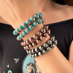 May include: Three beaded bracelets in teal and copper, copper, and dark grey. The bracelets are made of round beads and are stacked on a wrist. The bracelets are a fashion accessory.