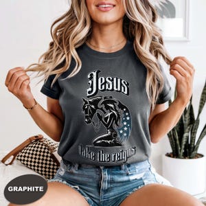 May include: Graphite-coloured t-shirt with the text "Jesus take the reigns" above an illustration of a horse and a woman in a cowboy hat. The design includes stars and a decorative font.