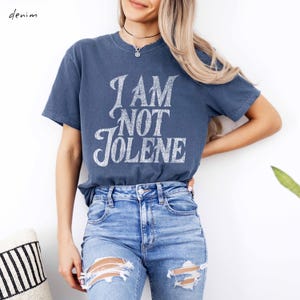 Pu&ograve; includere: Una maglietta blu denim con la scritta "I AM NOT JOLENE" in un carattere bianco invecchiato. La maglietta &egrave; abbinata a jeans strappati blu chiaro. La parola "denim" &egrave; nell'angolo in alto a sinistra.