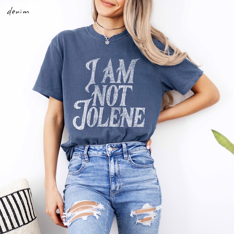 Pu&ograve; includere: Una maglietta blu denim con la scritta "I AM NOT JOLENE" in un carattere bianco invecchiato. La maglietta &egrave; abbinata a jeans strappati blu chiaro. La parola "denim" &egrave; nell'angolo in alto a sinistra.