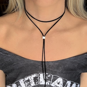 May include: A black choker necklace with a double-layered design. The necklace features a delicate, thin cord that forms a Y-shape, with a small, square silver-colored accent piece at the center. The necklace is worn around the neck.