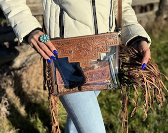 May include: A clear crossbody bag with a brown leather flap featuring intricate tooled floral designs. The bag has a long brown leather strap and brown fringe accents.