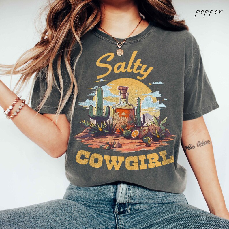 K&ouml;nnte beinhalten: Dunkelgraues T-Shirt mit einem W&uuml;sten-Themen-Grafik. Das Design zeigt eine Flasche, Kakteen und die Worte "Salty Cowgirl" in einem Vintage-Stil. Das Shirt hat kurze &Auml;rmel und einen Rundhalsausschnitt.