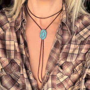 May include: A brown bolo tie with a turquoise stone pendant. The pendant is oval-shaped and surrounded by small turquoise stones. The tie is worn over a plaid shirt.