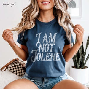 Pu&ograve; includere: Una maglietta blu navy con la scritta "I AM NOT JOLENE" in un carattere bianco invecchiato. La maglietta ha maniche corte e girocollo. La parola "midnight" &egrave; nell'angolo in alto a sinistra.