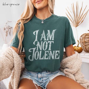 Pu&ograve; includere: Una maglietta verde acqua con la scritta "I AM NOT JOLENE" in un carattere bianco invecchiato. La maglietta &egrave; a maniche corte e ha un girocollo. Le parole "blue spruce" sono nell'angolo in alto a sinistra.
