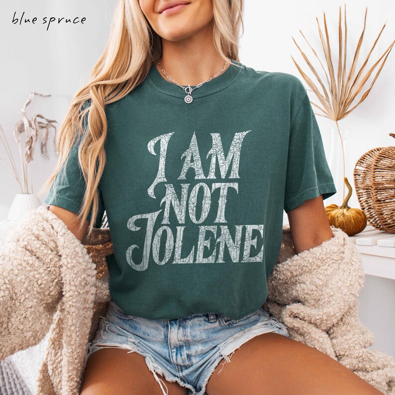 Pu&ograve; includere: Una maglietta verde acqua con la scritta "I AM NOT JOLENE" in un carattere bianco invecchiato. La maglietta &egrave; a maniche corte e ha un girocollo. Le parole "blue spruce" sono nell'angolo in alto a sinistra.
