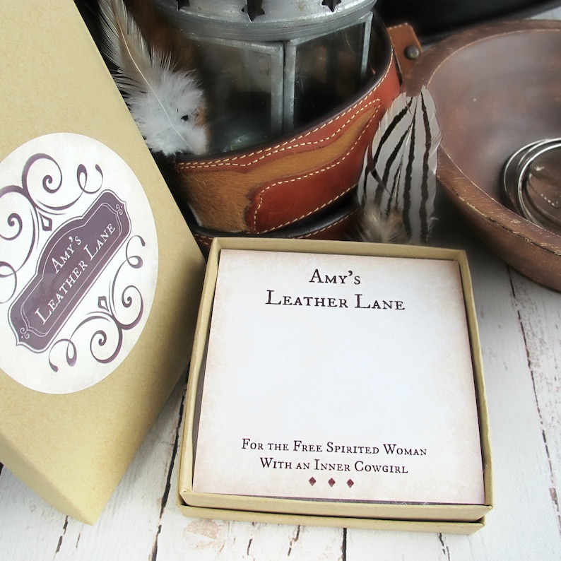 May include: A small, brown box with a white label that says "Amy's Leather Lane" and a decorative swirl design. The box is open and inside is a white card with the same text and the words "For the Free Spirited Woman With an Inner Cowgirl".