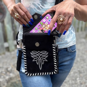 May include: Black crossbody bag with white stitching and a decorative embroidered design. The bag has a flap closure and a long strap. A smartphone is visible inside the bag.