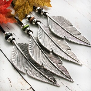 May include: Four silver feather pendants with black cords and decorative beads. The feathers have detailed veining and are arranged diagonally. The beads are silver, brown, and green. The background is a light wood-grain surface.