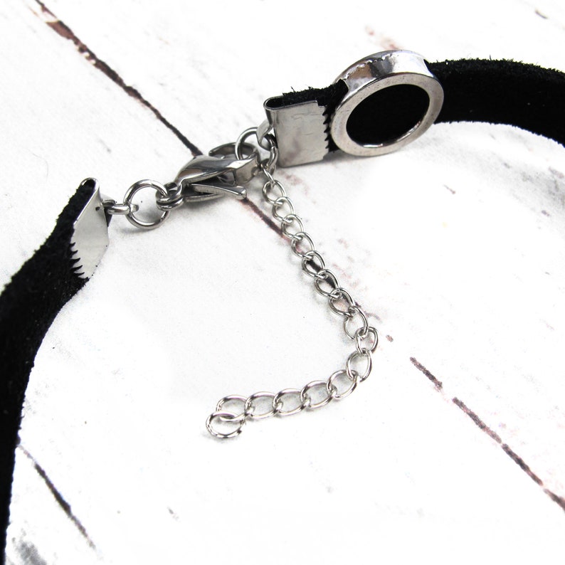 May include: A black suede choker necklace with a silver metal clasp and a round silver metal pendant. The pendant has a small hole in the centre.