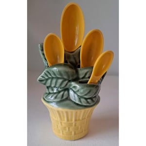 May include: A ceramic spoon holder with a basket weave design in yellow, and green leaves. The holder contains four yellow spoons. The spoons are oval shaped and appear to be made of plastic. The spoon holder is a decorative kitchen item.