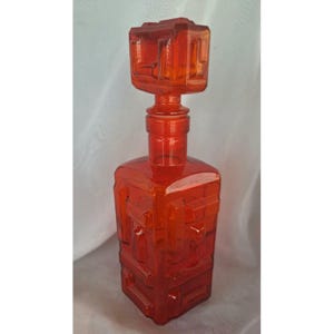 Puede incluir: Una jarra de vidrio rojo anaranjado vintage con un diseño geométrico. La jarra tiene una base cuadrada y un tapón a juego. El tapón tiene forma de cubo con un patrón geométrico. La jarra mide aproximadamente 30 cm de alto.
