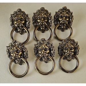 May include: A set of six antique brass-toned metal drawer pulls, each featuring a detailed lion head design with a circular ring handle suspended from the mouth. The hardware has a dark, weathered finish with visible highlights on the mane.