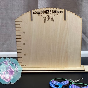 May include: Wooden earring display stand with a ruler on the side, labeled "NOLA BOOKS & BAUBLES". The stand has two slots for earrings. Two mermaid tail earrings are in the foreground.