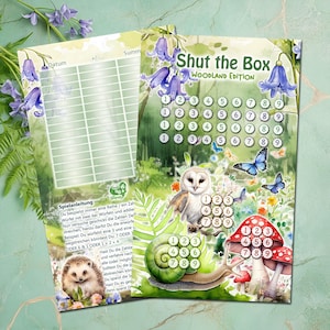 Op de afbeelding: Een groen en geel spelbord met de titel "Shut the Box Woodland Edition". Het bord heeft de nummers 1 tot en met 9 in rijen. Er zijn illustraties van een slak, een uil, een paddenstoel en vlinders. Het spelbord is voor een dobbelspel.