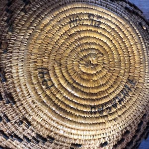 May include: Close-up of a handwoven basket with a spiral design. The basket is made of natural-colored fibers with dark brown accents. The circular pattern creates a textured surface, showcasing the craftsmanship of the basket.