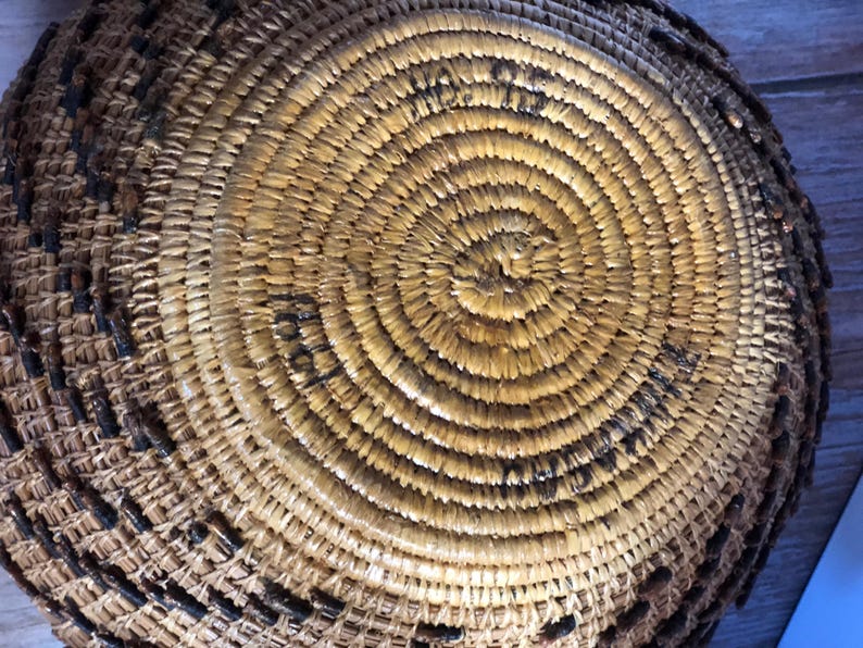 May include: Close-up of a handwoven basket with a spiral design. The basket is made of natural-colored fibers with dark brown accents. The circular pattern creates a textured surface, showcasing the craftsmanship of the basket.
