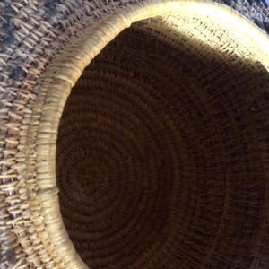 May include: A woven basket with a circular opening. The basket is made of natural fibers, with a light-colored rim and a dark brown interior. The exterior features a pattern of dark and light woven sections.