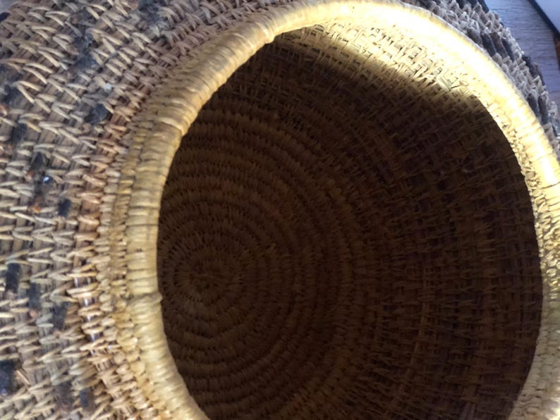 May include: A woven basket with a circular opening. The basket is made of natural fibers, with a light-colored rim and a dark brown interior. The exterior features a pattern of dark and light woven sections.