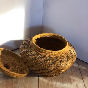 May include: A handwoven basket with a lid, crafted from natural fibers. The basket features a round shape with a wide opening and a lid that sits on top. The exterior is adorned with a repeating pattern of dark brown accents.