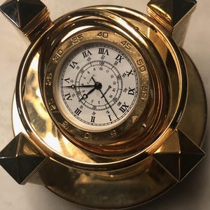 May include: A gold-toned desk clock featuring a circular face with Roman numerals and a secondary inner ring with Arabic numerals. The clock is encased in a rotating gold-colored frame with four pyramid-shaped corner accents on a circular base.