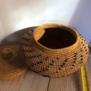 May include: A handwoven basket with a lid, crafted from natural fibers. The basket is round with a wide opening and a lid. Dark accents are woven into the sides. The basket is sitting on a wooden surface.