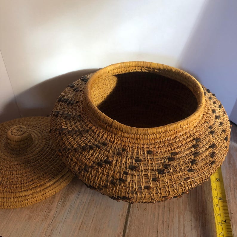 May include: A handwoven basket with a lid, crafted from natural fibers. The basket is round with a wide opening and a lid. Dark accents are woven into the sides. The basket is sitting on a wooden surface.