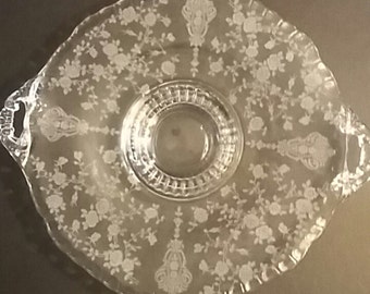 May include: A clear glass serving platter with two side handles and a scalloped edge. The surface features an etched floral pattern of roses and vines, with a central circular section surrounded by a decorative ribbed border design.