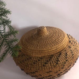 May include: A woven basket with a lid, featuring a natural tan color with dark brown accents. The basket has a round shape and a textured surface. The lid has a knob on top for easy opening. The basket is likely handmade.