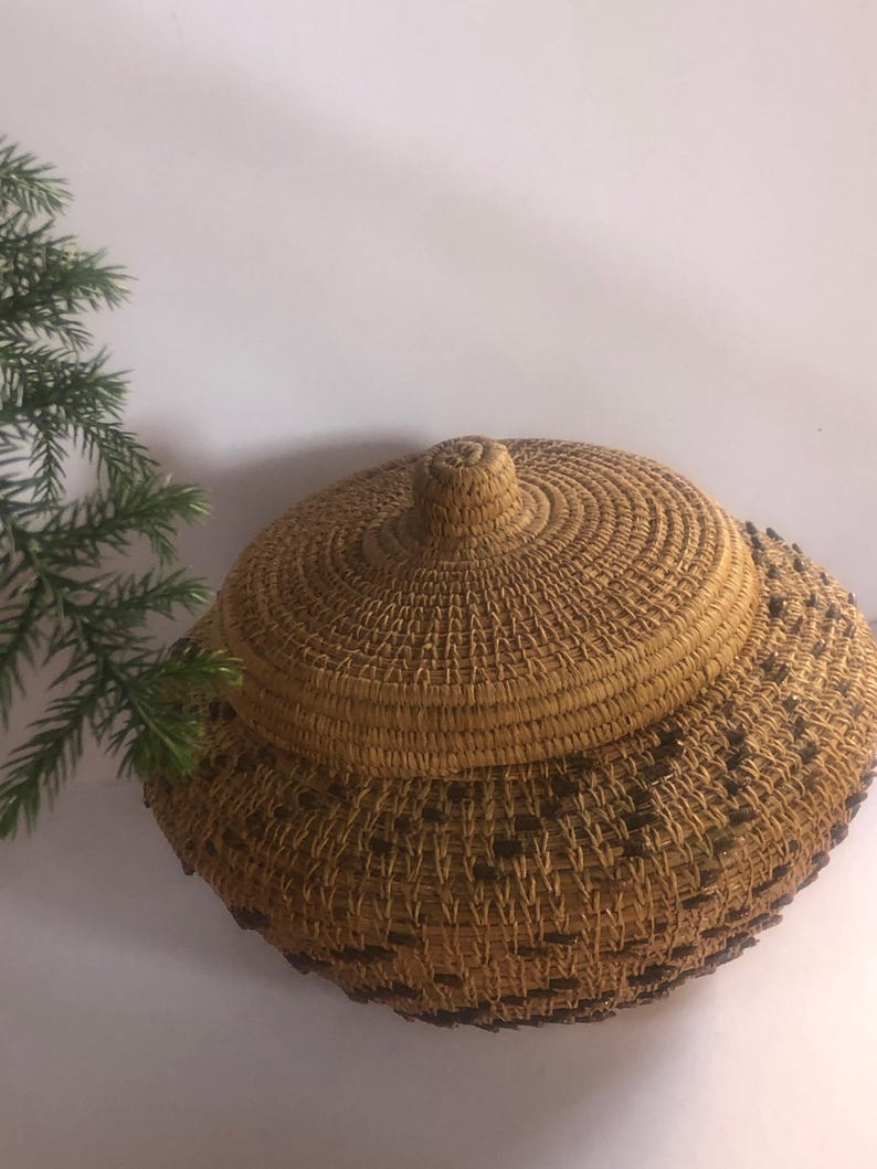 May include: A woven basket with a lid, featuring a natural tan color with dark brown accents. The basket has a round shape and a textured surface. The lid has a knob on top for easy opening. The basket is likely handmade.