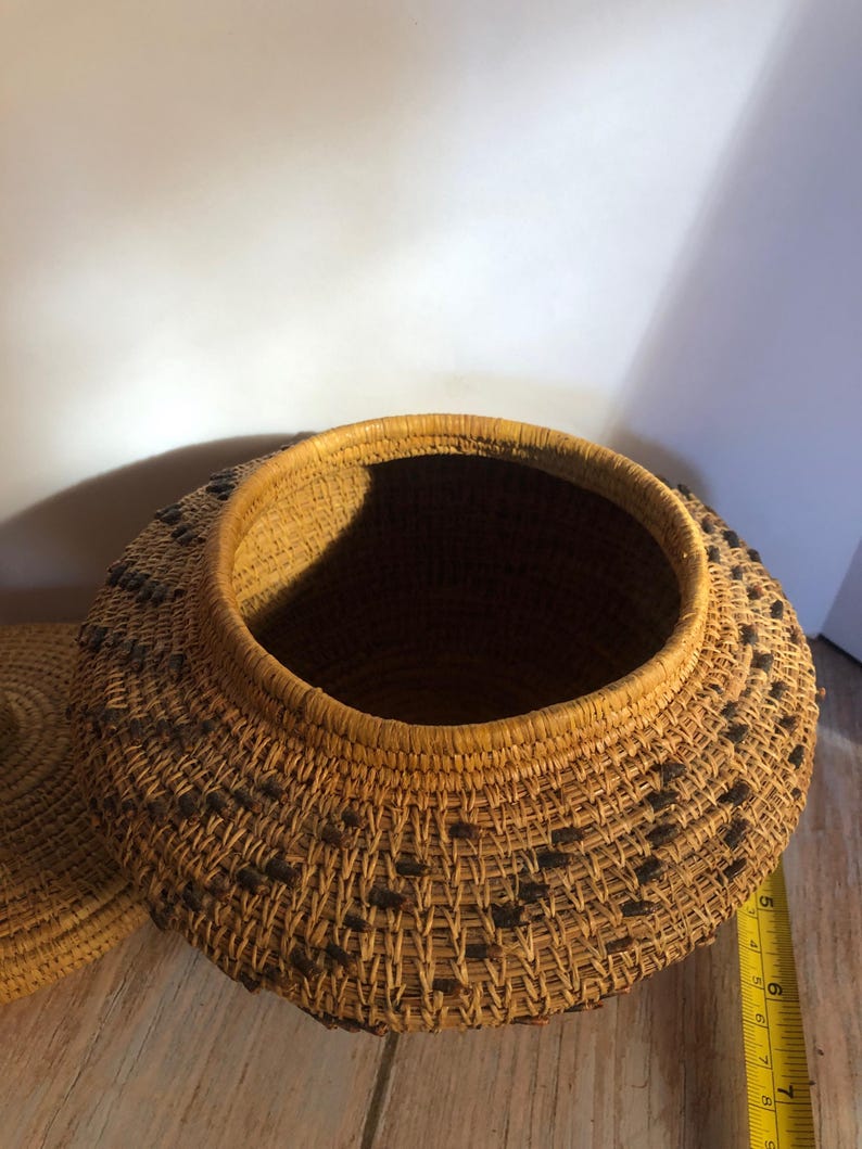 May include: A round, woven basket with a lid. The basket is made of natural-colored fibers with a darker pattern of small, square accents. The rim and lid edge are reinforced with a darker, thicker weave. The basket is approximately 8 inches in diameter.
