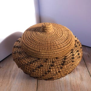 May include: A round, lidded basket made of woven natural fibers. The basket has a light brown color with darker brown accents in a repeating pattern around the base. The lid features a small knob and concentric circles of woven material.