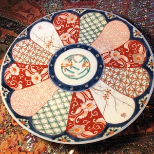 May include: A decorative ceramic plate with a blue rim and a central circular design. The plate features a variety of patterned sections in red, white, and green, with floral and geometric motifs. The center circle has a floral design.